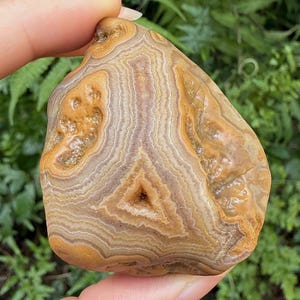 May include: A polished agate stone featuring concentric bands of tan, beige, and brown colors. The surface displays a natural triangular cavity in the center and textured, crystalline formations along the sides of the stone's layered structure.