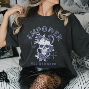 Peut inclure: Un t-shirt gris foncé avec un motif de crâne et de serpent et le texte "EMPOWER ALL GENDERS".
