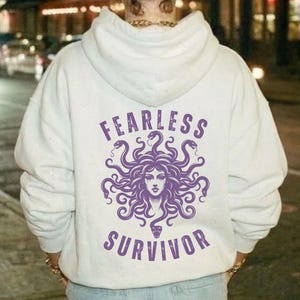 May include: A white hoodie with a purple Medusa graphic and the words "FEARLESS" and "SURVIVOR" printed on the back. The design features a detailed illustration of Medusa with snakes for hair.