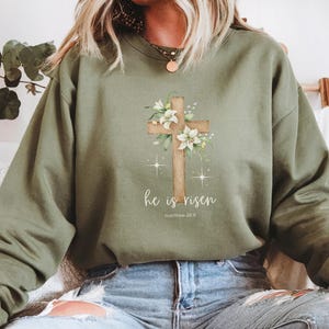 Op de afbeelding: Een groen sweatshirt met een bloemig kruisontwerp en de tekst "Hij is opgestaan" met de bijbelverwijzing "Mattheüs 28:6".