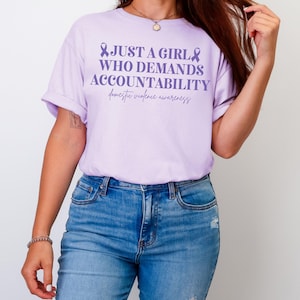 May include: A light purple t-shirt with the text "JUST A GIRL WHO DEMANDS ACCOUNTABILITY" and "domestic violence awareness" in a script font. The shirt is paired with blue jeans.