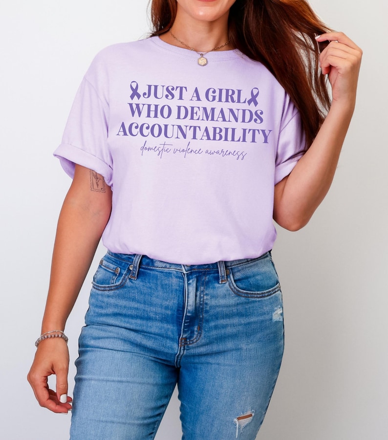 May include: A light purple t-shirt with the text "JUST A GIRL WHO DEMANDS ACCOUNTABILITY" and "domestic violence awareness" in a script font. The shirt is paired with blue jeans.