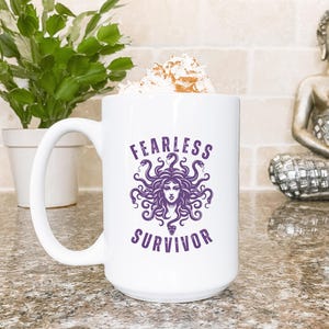 Könnte beinhalten: Eine weiße Keramiktasse mit großem Henkel, mit den lila Worten "FEARLESS SURVIVOR" und einer Medusa-Illustration. Die Tasse ist mit einem Schlagsahne-Topping mit Zimt bestreut gefüllt.