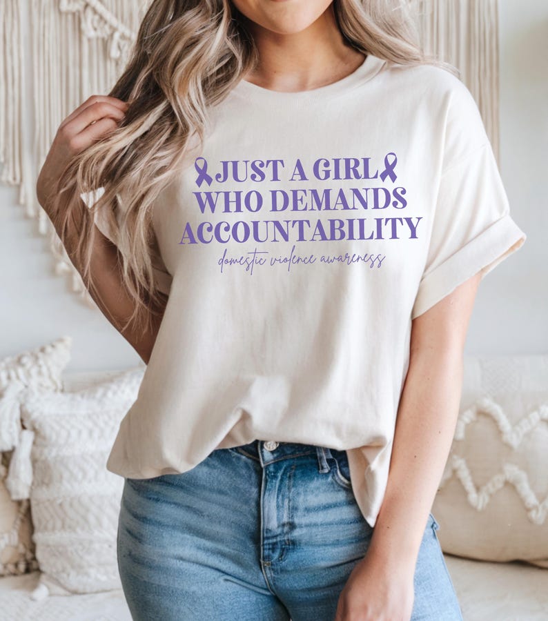 May include: Cream-colored t-shirt with the text "JUST A GIRL WHO DEMANDS ACCOUNTABILITY" in purple, along with a purple ribbon and the words "domestic violence awareness". The shirt is paired with blue jeans.