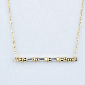 May include: A delicate gold-colored necklace with a bar pendant. The pendant features a series of small, gold-colored beads interspersed with silver-colored beads. The necklace is on a white background. The text "Rise n Shine" is in the lower right corner.