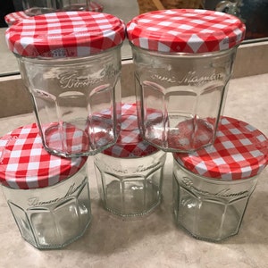 May include: Five clear glass jars with red and white checkered lids. The jars have a hexagonal shape and the words "Bonne Maman" are embossed on the front. The lids have a white border and are made of fabric.