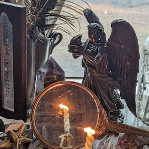 Peut inclure: Un autel sur une table avec divers objets, dont une petite figurine d'ange, une bougie, un miroir, un bocal et plusieurs cercles en bois avec des symboles écrits dessus. L'autel est décoré de pétales de fleurs séchées et de cristaux.