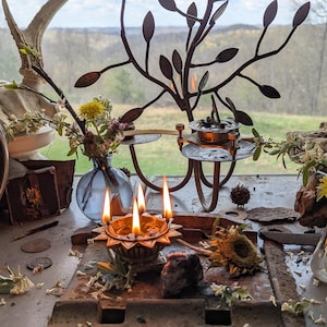 Può includere: Una natura morta rustica con un candelabro in metallo con candele accese, un piccolo vaso di vetro con fiori, una scultura di albero in metallo e un ramo di legno con un teschio. La scena è ambientata su un tavolo di legno con una finestra sullo sfondo.