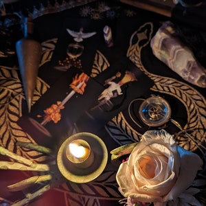 May include: A dark, mystical scene with a lit candle, a white rose, and tarot cards on a black and gold patterned cloth. There are also other objects, such as a compass and a crystal, scattered around the scene.