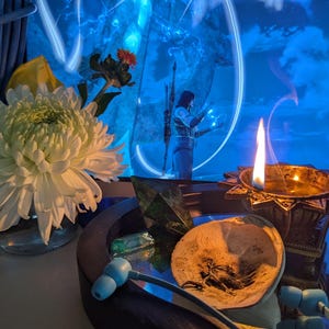 May include: A still life arrangement featuring a white chrysanthemum in a glass vase, a lit candle in a decorative holder, and a small bronze statue. The background is a blue-toned scene with a figure. Other items include a seashell and blue earbuds.