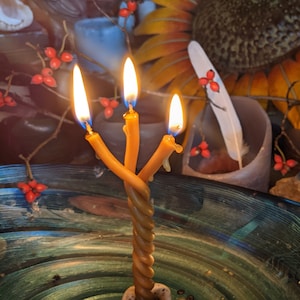 Pode incluir: Uma vela de três pavios com um corpo de cera marrom torcido está acesa e queimando. A vela é colocada em uma tigela com um padrão de redemoinho verde e azul. A vela é cercada por outros objetos, incluindo um girassol, uma pena e um cristal.