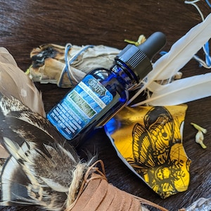 以下が含まれることがあります： ドロップトップ付きの青いガラス瓶で、「グレイオウルスピリッツ魔法のブレンドオイルプロテクション」とラベル付けされています。羽根、お香、黒い蝶の模様が描かれた金箔で囲まれています。