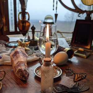 Puede incluir: Una mesa con una variedad de objetos, incluyendo una vela encendida, un vaso de agua, un cristal, una pequeña botella, una brújula, una figurita de pájaro, un huevo blanco y alas de mariposa. Los objetos están dispuestos en círculo sobre una mesa de madera.