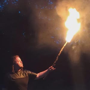 May include: A person holding a lit torch in a dark outdoor setting. The torch emits bright orange and yellow flames, with smoke trailing upwards. The person is wearing a dark-colored top and pants, standing in a grassy area with trees in the background.