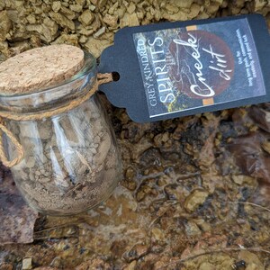 May include: A small glass jar with a cork lid filled with brown dirt. The jar is tied with twine and has a black tag attached that reads "Grey Kindred Spirits Creek Dirt".