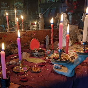 以下が含まれることがあります： 紫、ピンク、白のキャンドルマジックの祭壇で、さまざまなクリスタル、小さな赤いハート型の箱、ローリエの葉が置かれています。祭壇は、白いレースの縁取りのある濃い紫色の布で覆われています。キャンドルに火が灯され、背景にはいくつかの彫像があります。