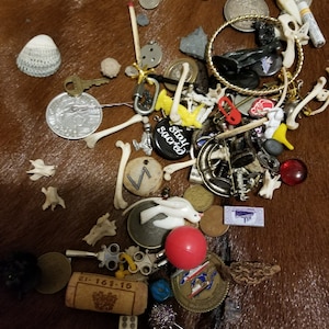 May include: A collection of assorted objects including coins, bones, charms, a seashell, and a cork. The items are scattered on a brown, textured surface. Notable items include a key, a red ball, and a charm with the words "stay sacred".