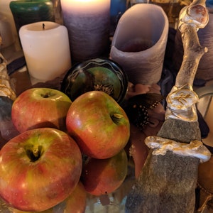 May include: A still life arrangement with three red apples, two candles, a small bowl, and a wooden wand with gold accents. The apples are arranged in the center of the image, with the candles and wand surrounding them.