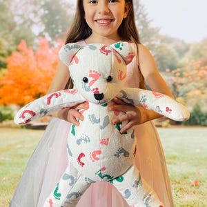May include: A white teddy bear with a colorful footprint pattern. The bear is being held by a young girl wearing a pink dress.