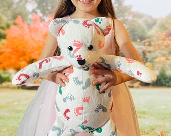 May include: A white teddy bear with a colorful footprint pattern. The bear is being held by a young girl wearing a pink dress.