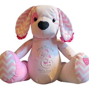 May include: Pink and white patchwork dog plush toy with embroidered details. The dog has a pink and white chevron pattern on its ears, legs, and feet. The dog's body is pink with white stripes and has an embroidered seahorse with the text "Love the Sea" and "Santa Cruz California".
