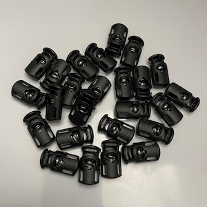 May include: A collection of black cord locks with a spring mechanism. These plastic cord locks are designed to secure and adjust cords, ropes, or drawstrings. The image shows a close-up view of the cord locks, highlighting their design and functionality.