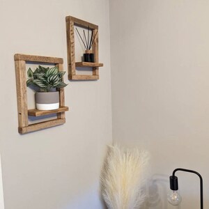May include: Two rustic wooden shelves with a plant in a gray pot on one shelf and a black diffuser on the other shelf. The shelves are mounted on a white wall.