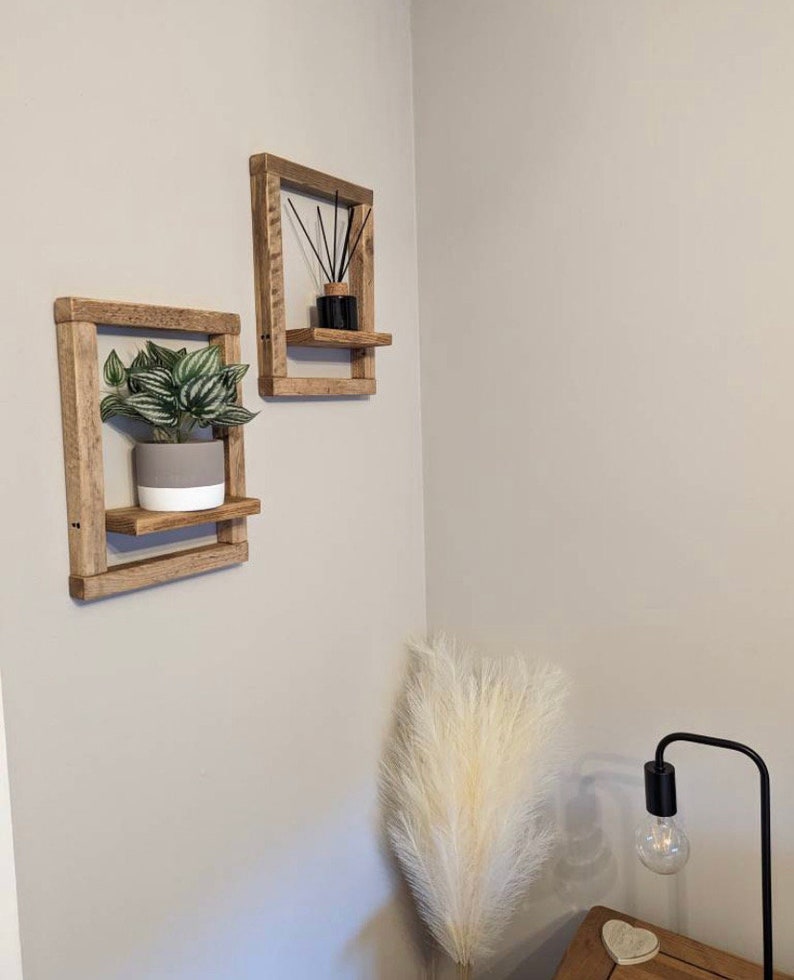 May include: Two rustic wooden shelves with a plant in a gray pot on one shelf and a black diffuser on the other shelf. The shelves are mounted on a white wall.