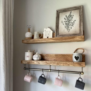 May include: Rustic wooden shelves with a metal rail for hanging mugs. The shelves are decorated with a ceramic house sign that reads "There's no place like home", a ceramic jar, a ceramic garlic, and a framed botanical print. The mugs hanging on the rail are white, black, pink, and gray.