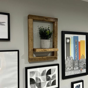 May include: A rustic wooden shelf with a single shelf and a small potted plant. The shelf is mounted on a wall with black framed pictures on either side.
