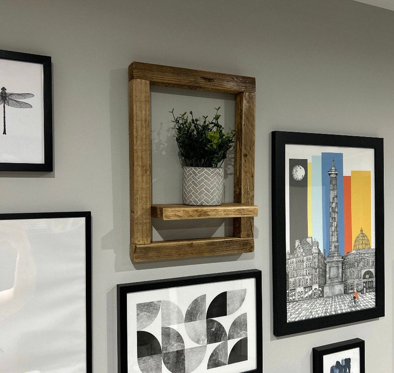 May include: A rustic wooden shelf with a single shelf and a small potted plant. The shelf is mounted on a wall with black framed pictures on either side.