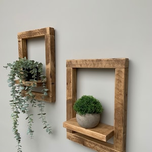 May include: Two rustic wooden shelves with a square frame and a small shelf in the front. The shelves are mounted on a white wall. A small potted plant sits on the top shelf of the left shelf and a small potted plant sits on the shelf of the right shelf.