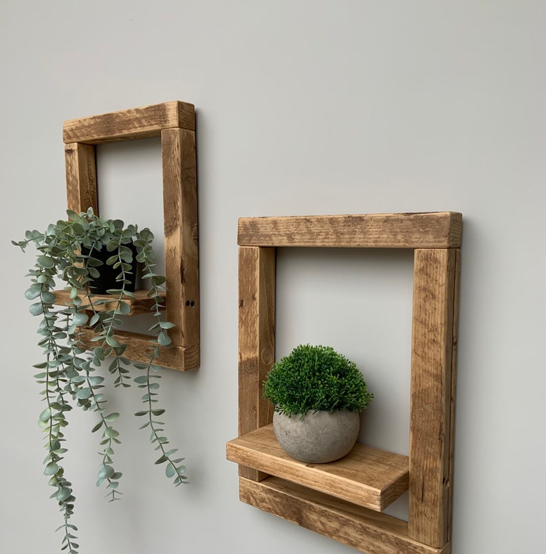 May include: Two rustic wooden shelves with a square frame and a small shelf in the front. The shelves are mounted on a white wall. A small potted plant sits on the top shelf of the left shelf and a small potted plant sits on the shelf of the right shelf.