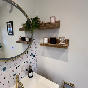 May include: Two rustic wooden shelves with a white background. The top shelf has a pink candle and a small figurine. The bottom shelf has a clear glass jar with cotton swabs, a black jar, and a clear glass jar with cotton balls.