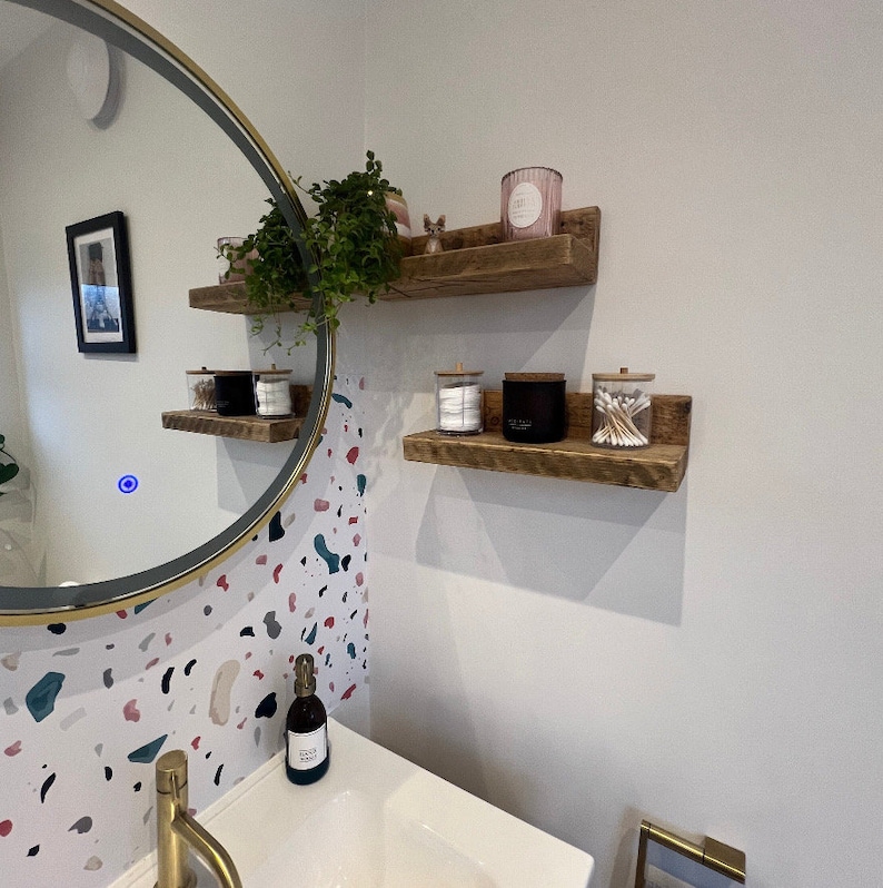 May include: Two rustic wooden shelves with a white background. The top shelf has a pink candle and a small figurine. The bottom shelf has a clear glass jar with cotton swabs, a black jar, and a clear glass jar with cotton balls.