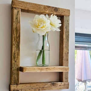 May include: A rustic wooden shelf with two shelves, featuring a clear glass vase with white flowers. The shelf is mounted on a white wall.