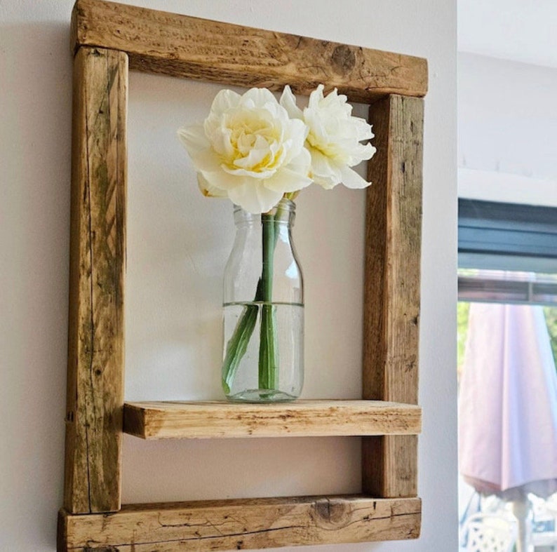 May include: A rustic wooden shelf with two shelves, featuring a clear glass vase with white flowers. The shelf is mounted on a white wall.