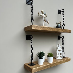 May include: Two wooden shelves with black metal chains hanging from the wall. The shelves are decorated with small decorative items, including a house, plants, and fish.