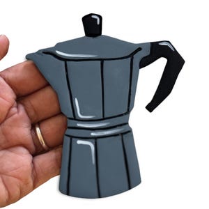 May include: A gray coffee maker with a black handle and top. The coffee maker is a classic Italian design, with a rounded base and a straight, vertical body. The handle is black and curved. The coffee maker is held by a hand.
