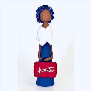 May include: A handcrafted figurine of a person with blue hair curlers, a blue dress, and a white shawl. The figurine holds a red bag with the name "Juanita" in white script. The figure has a brown face and is set against a white background.