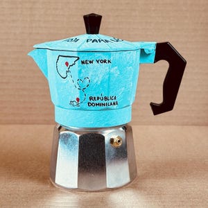 May include: A light blue stovetop espresso maker with a black handle and lid. The maker has a map design with the words "NEW YORK" and "REPUBLICA DOMINICANA" printed on it. The base is silver.