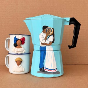 Op de afbeelding: Een lichtblauwe, beschilderde espressomaker en twee bijpassende emaille mokken. De espressomaker heeft een geschilderd stel dat elkaar omhelst. De mokken hebben geschilderde figuren in traditionele kleding.