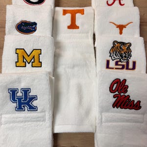 Peut inclure: Une collection de serviettes en tissu éponge blanc, chacune ornée de logos universitaires brodés, notamment le G de Georgia, le Gator de Floride, le M de Michigan, le UK de Kentucky, le T de Tennessee, le A d'Alabama, le Texas Longhorn, le tigre LSU et le logo Ole Miss.