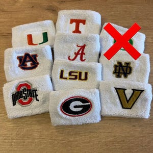 Puede incluir: Una colección de muñequeras de felpa blancas, cada una con un logotipo deportivo universitario bordado diferente. Los logotipos incluyen U, T, A, Auburn, LSU, Ohio State, G, Notre Dame y V. Una muñequera está marcada con una gran X roja.