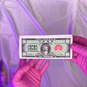 Könnte beinhalten: Ein Stapel Hell Bank Notes, jede mit einem Porträt, chinesischen Schriftzeichen und dem Text "TEN THOUSAND DOLLARS". Die Noten haben eine grüne und rote Farbgebung mit aufwendigen Designs. Die Noten werden von einer Person mit rosa Handschuhen gehalten.