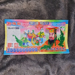 Pode incluir: Uma nota de papel retangular com uma ilustração colorida de uma figura em trajes tradicionais, crianças puxando um carrinho e o texto "the hell bank corporation" junto com "10000000000" e "YA-H11288" em um fundo vibrante e multicolorido.