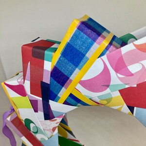Pu&ograve; includere: Una ghirlanda di carta colorata composta da pi&ugrave; pezzi di carta a motivi, ognuno con un design diverso. La ghirlanda &egrave; decorata con nastro giallo e un nastro viola.