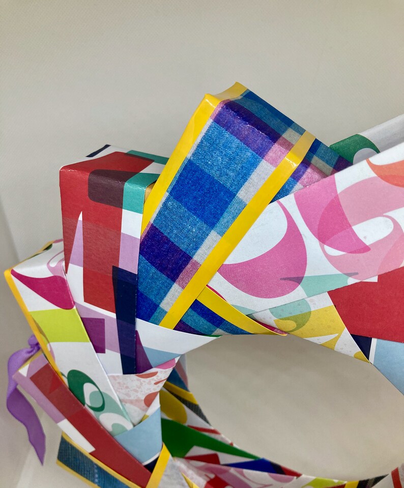 Pu&ograve; includere: Una ghirlanda di carta colorata composta da pi&ugrave; pezzi di carta a motivi, ognuno con un design diverso. La ghirlanda &egrave; decorata con nastro giallo e un nastro viola.
