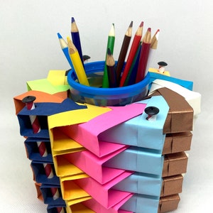 Pu&ograve; includere: Un porta matite in origami colorato realizzato con carta piegata in tonalit&agrave; di blu, giallo, rosa, arancione e marrone. Il porta matite ha un bicchiere di plastica trasparente all'interno per contenere le matite. Il porta matite &egrave; decorato con piccole viti nere. Ci sono matite colorate nel bicchiere, tra cui rosso, blu, verde e marrone.