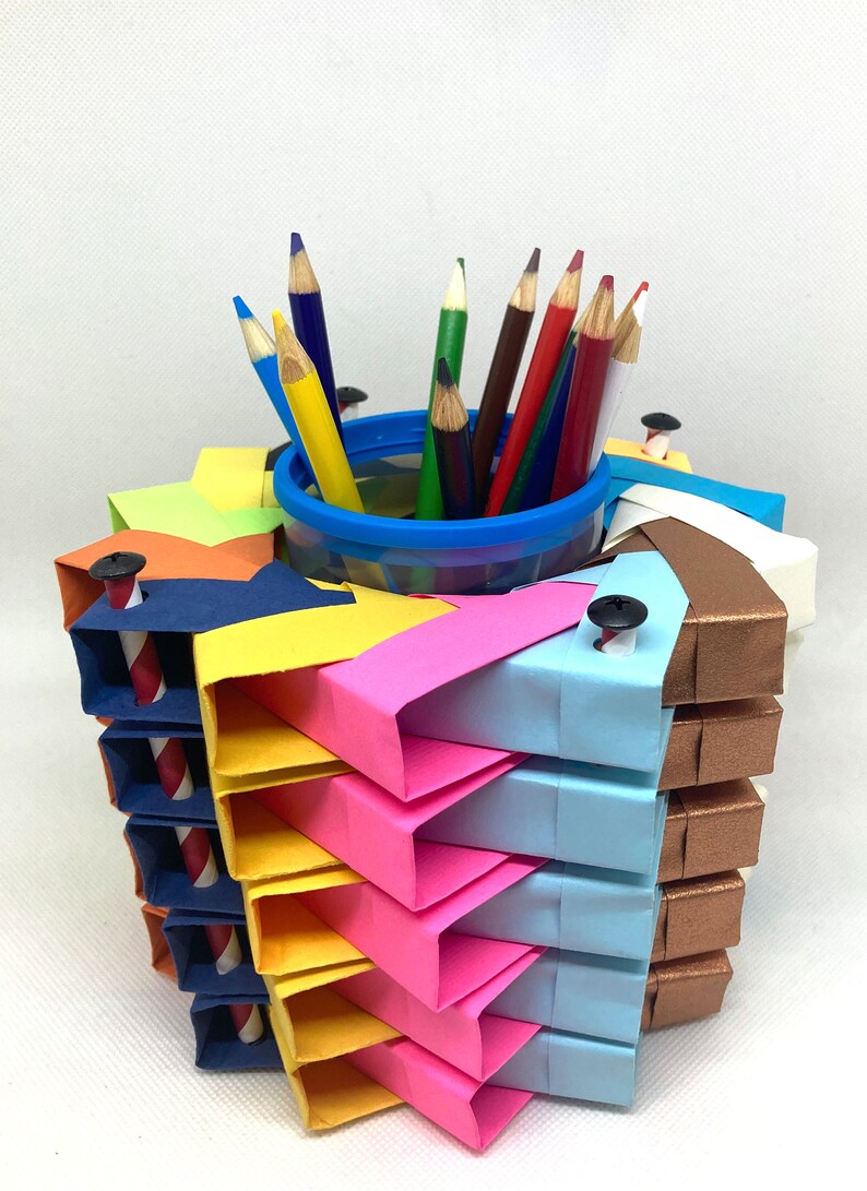 Pu&ograve; includere: Un porta matite in origami colorato realizzato con carta piegata in tonalit&agrave; di blu, giallo, rosa, arancione e marrone. Il porta matite ha un bicchiere di plastica trasparente all'interno per contenere le matite. Il porta matite &egrave; decorato con piccole viti nere. Ci sono matite colorate nel bicchiere, tra cui rosso, blu, verde e marrone.